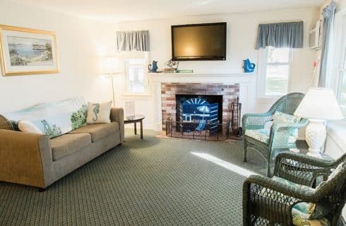 Living room of a cottage with fireplace, TV, tan couch and two green wicker chairs in front of a large window