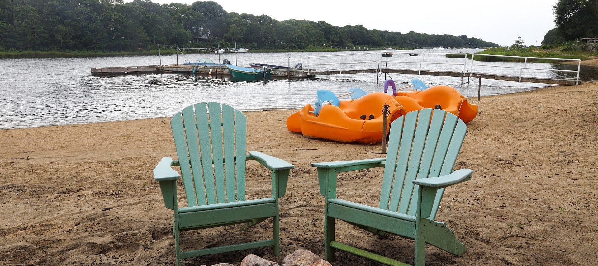 Two green adirondack chairs on a beach with paddle boats and a long dock