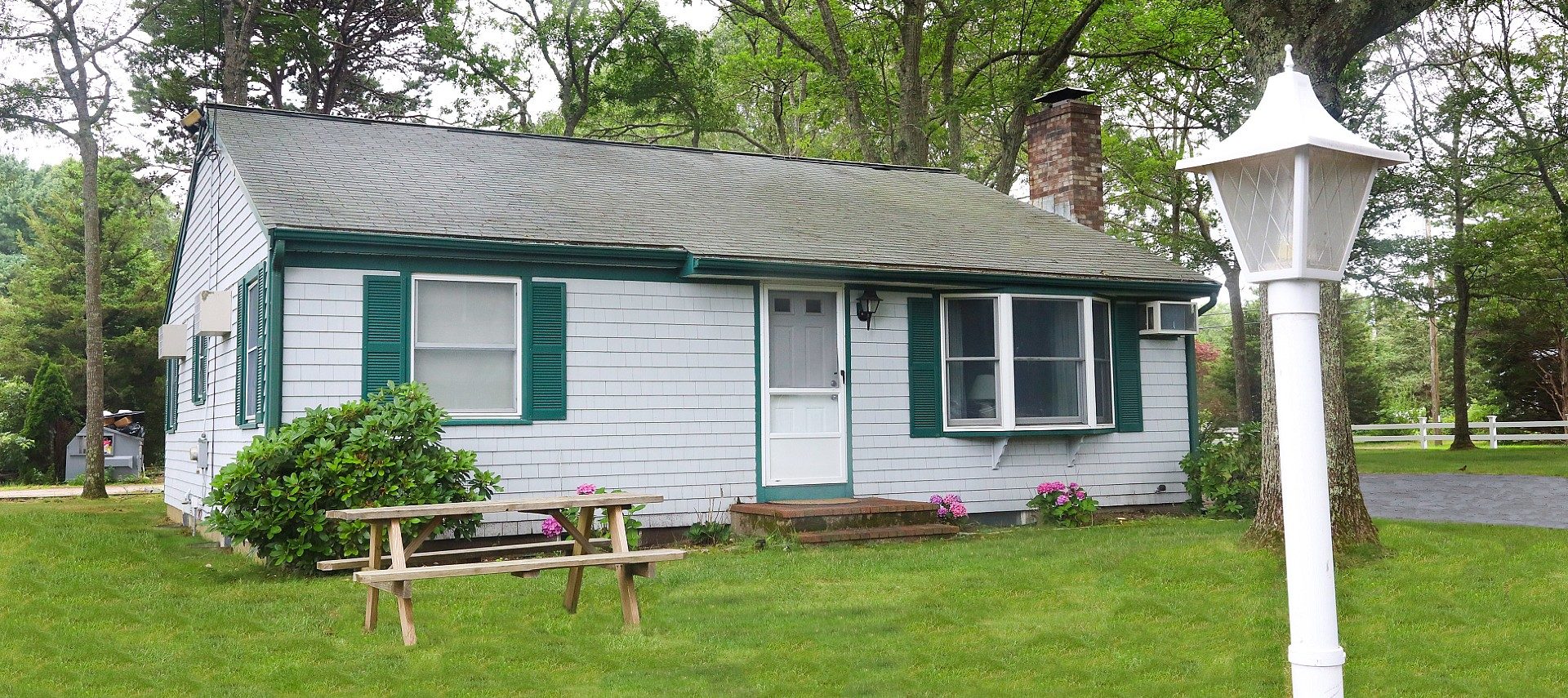 Front facade of a cottage with grey siding and green shutters and white lamppost